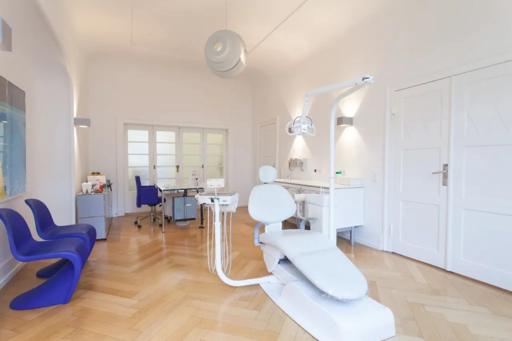 Helles Behandlungszimmer mit Behandlungsstuhl, Fischgrätparkett und blauen Stühlen