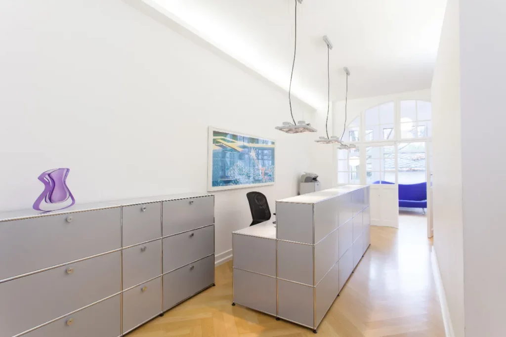 Empfangsbereich mit Tresen und grauen Sideboards, im Hintergrund das Wartezimmer