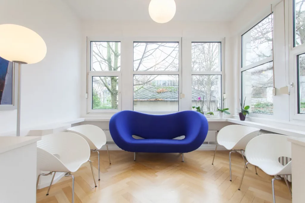 Wartezimmer mit blauem Sofa, weißen Stühlen und großen Fenstern mit Blick ins Grüne